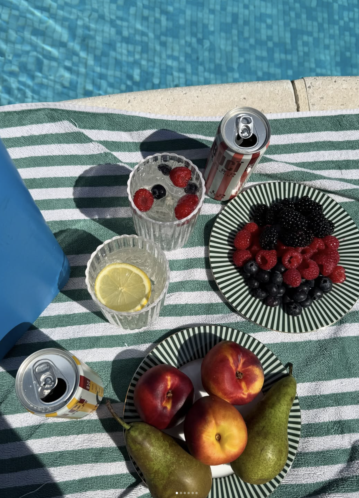 Two cans of BUCI hard kombucha by a pool next to snacks on a towel.
